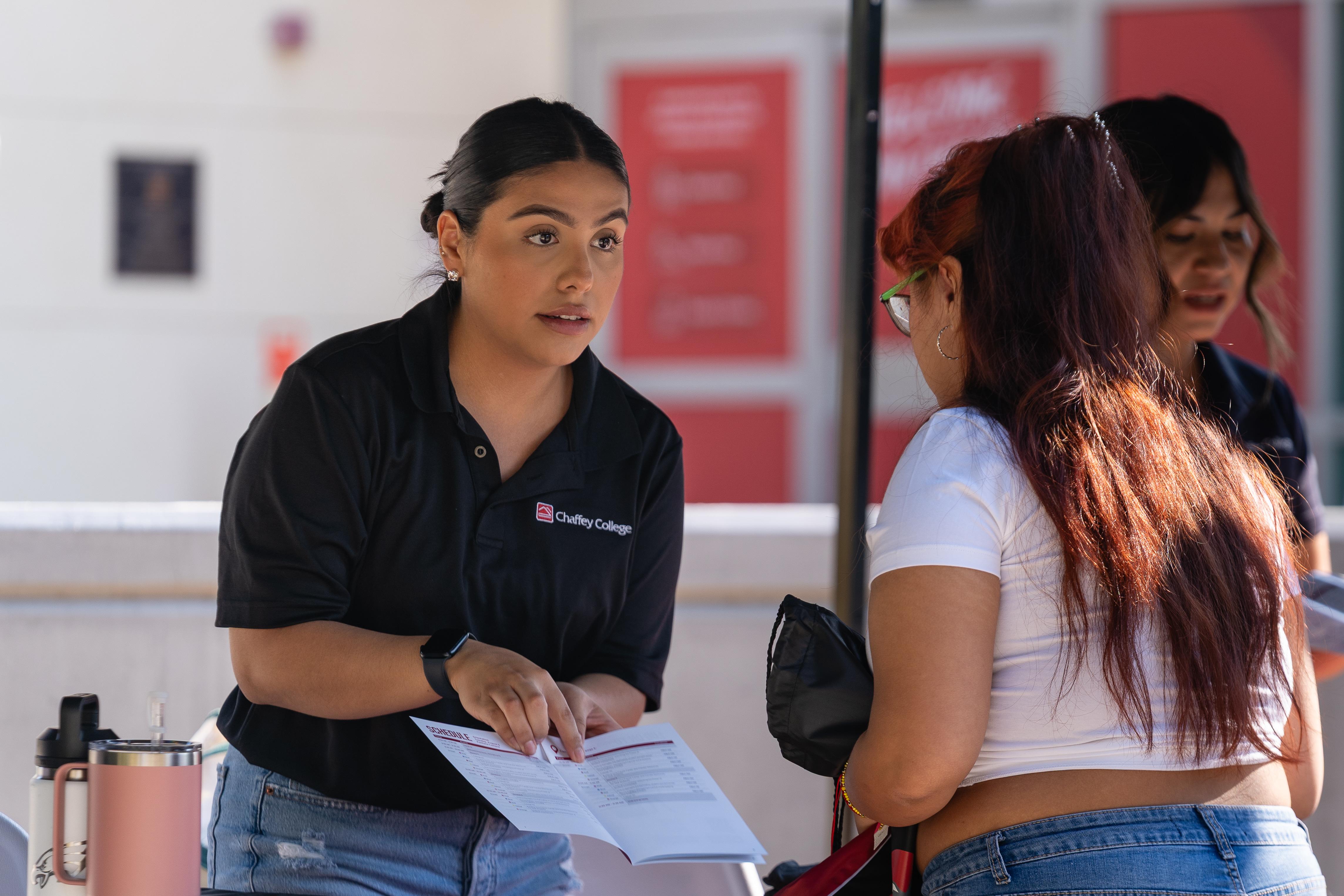 A Chaffey College representative in a black polo shirt points at a brochure while speaking to a student.
