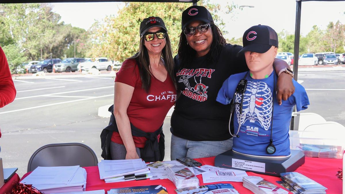 Two smiling women wearing Chaffey shirts and caps stand together at an outdoor table covered with brochures and papers. A medical training mannequin, also wearing a cap, is displayed beside them under a canopy, with parked cars visible in the background.