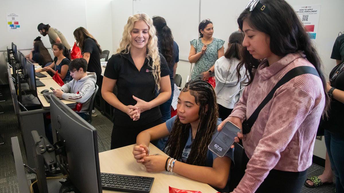 A classroom scene with a group of people engaged in a computer activity. A woman with long blonde hair in a black shirt is smiling and interacting with a seated young woman at a computer. Another woman stands nearby holding a phone. Several other individuals are seated at computers or standing in the background, creating a lively and collaborative atmosphere.