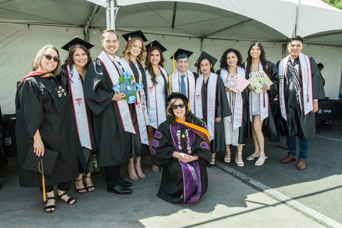 A group of smiling graduates in caps and gowns, with ten standing and one kneeling. Many wear stoles with a medical symbol, and one holds a sign that reads TCU LATER