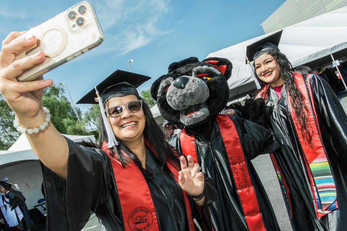 A woman in a graduation cap and gown takes a selfie with a panther mascot and another graduate. Their red stoles read, CHAFFEY COLLEGE FOUNDED IN 1883 and CHAFFEY COLLEGE