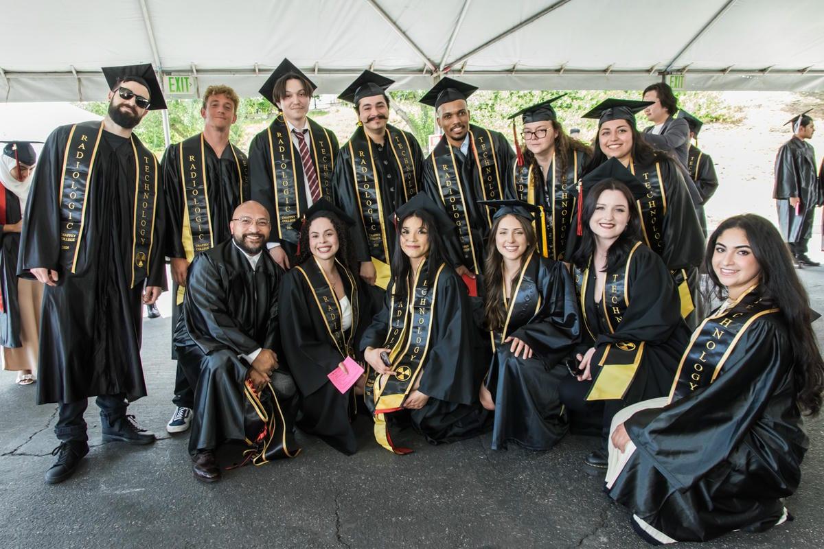 A group of smiling graduates in black caps and gowns with gold stoles, many reading RADIOLOGIC TECHNOLOGY, pose together under a tent. They celebrate their academic achievement.