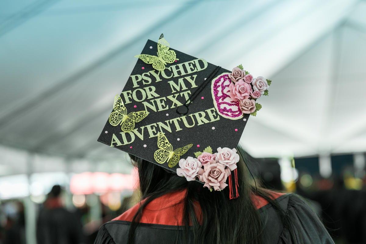 A decorated black graduation cap reads, PSYCHED FOR MY NEXT ADVENTURE. It features gold butterflies, a pink brain, and roses, worn by a graduate at a ceremony.