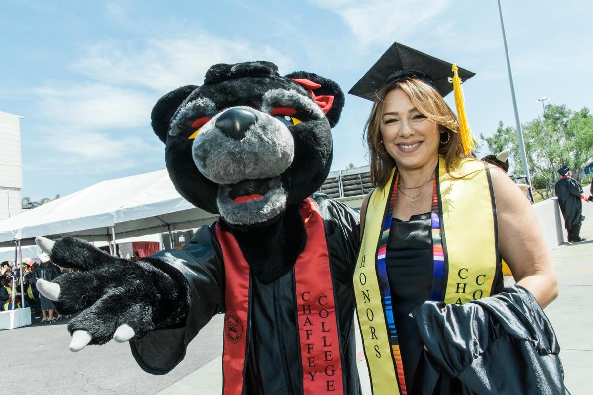 A graduate smiles with the Chaffey College panther mascot, both in academic regalia. Stoles read HONORS and CHAFFEY COLLEGE