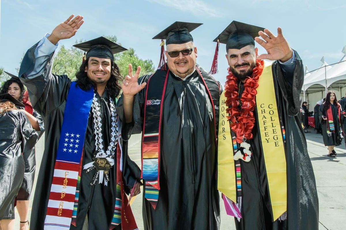 Three smiling graduates in caps and gowns wave at their outdoor ceremony. Their stoles read CHAFFEY COLLEGE, HONORS, and CLASS OF 2023.