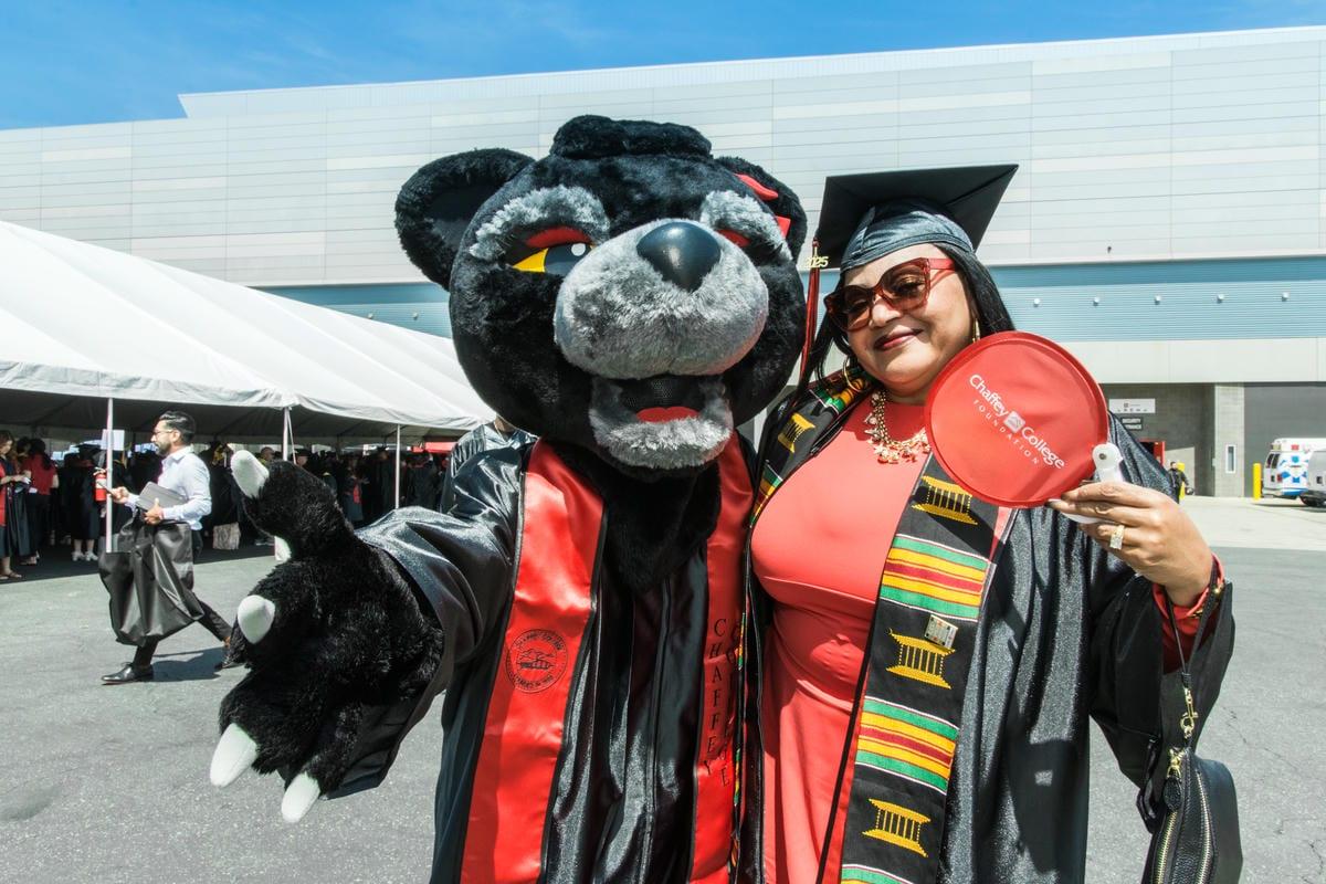 A graduate in cap and gown smiles with the Chaffey College panther mascot. She holds a red fan that reads Chaffey College Foundation.
