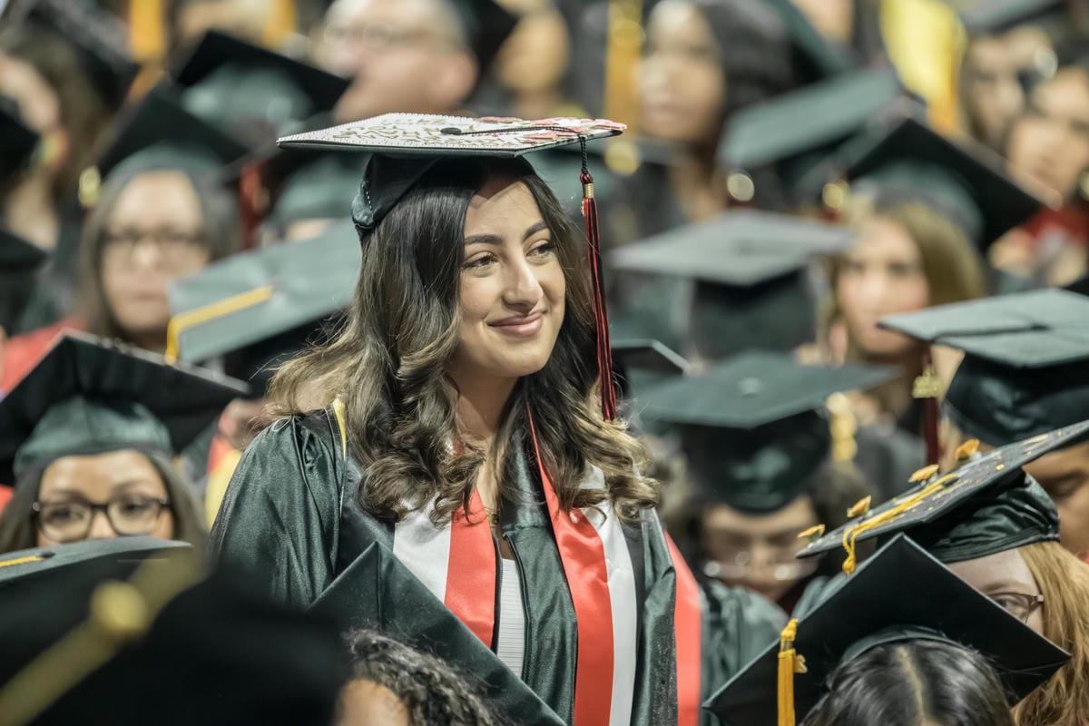 A smiling graduate in a black gown, red and white stole, and decorated cap stands amidst a crowd of fellow graduates.