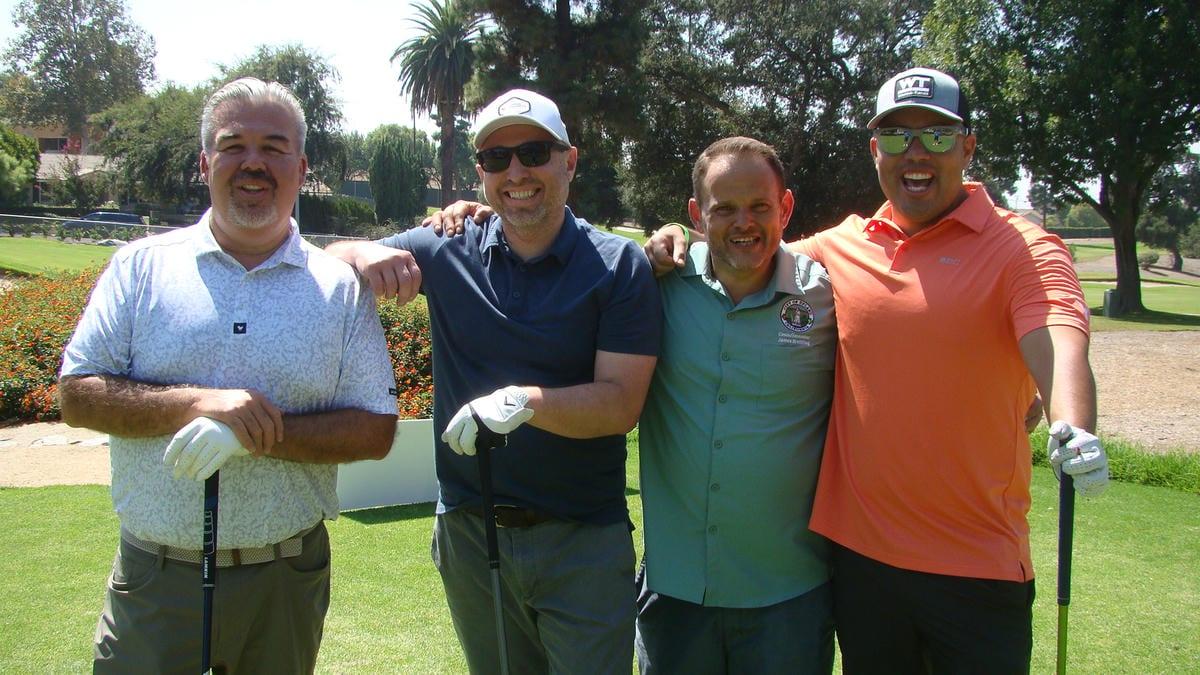 Four smiling men in golf attire pose on a sunny golf course with their clubs. They appear to be enjoying a golf outing together.