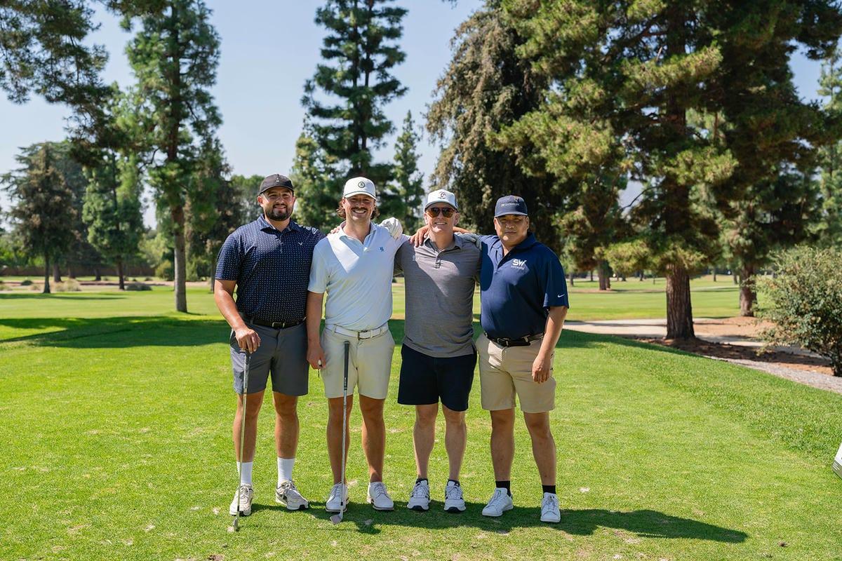 Four golfers stand arm in arm with golf clubs on a sunny, tree-lined course.