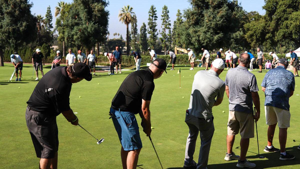 A group of men practice putting on a sunny golf green. Several men in the foreground are bent over, holding putters, surrounded by golf balls.