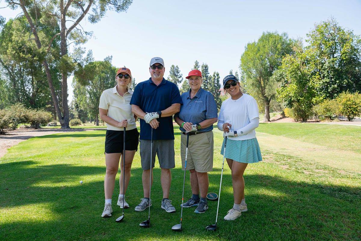 Four smiling golfers pose with their clubs on a green fairway surrounded by trees.