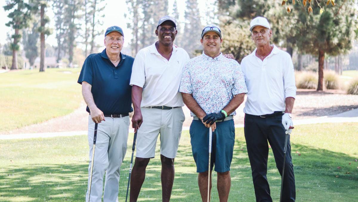 Four golfers smile while holding clubs on a sunny course with trees in the background, dressed in casual golf attire.