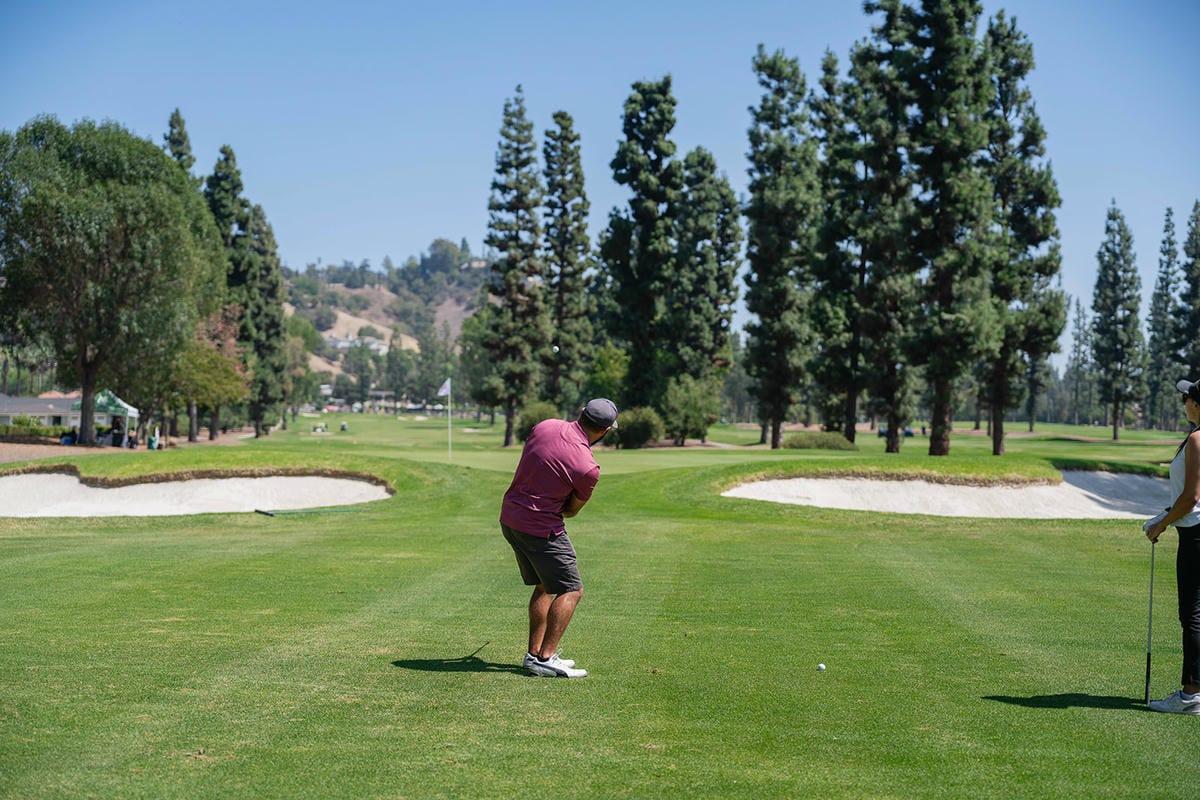 A golfer takes an approach shot toward a green framed by sand bunkers and tall pine trees.
