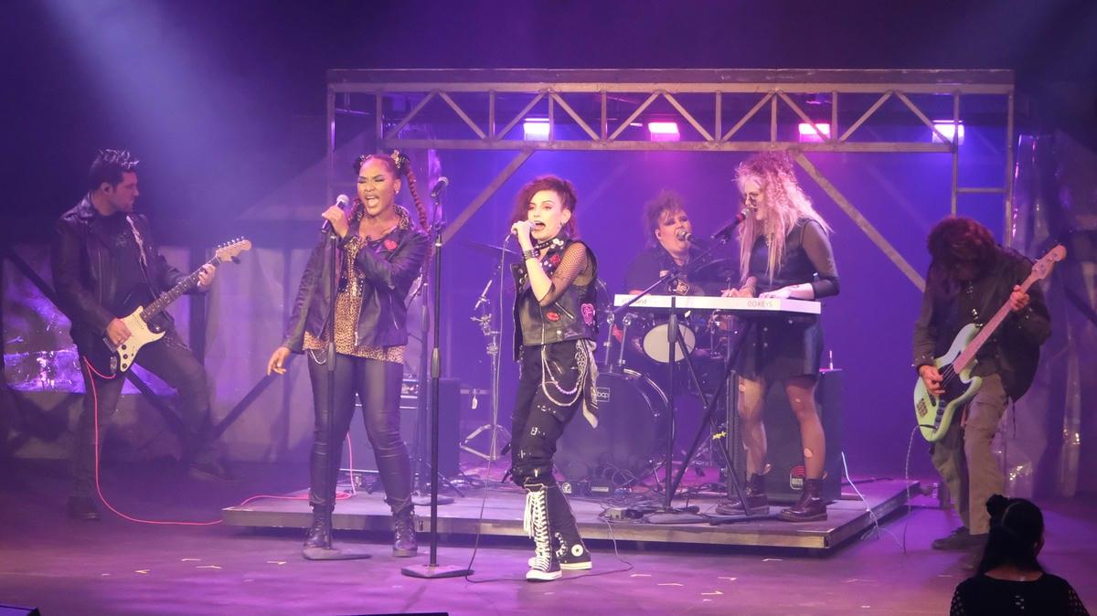 Music group performing live on a stage with microphones, guitars, drums, and colorful stage lighting.