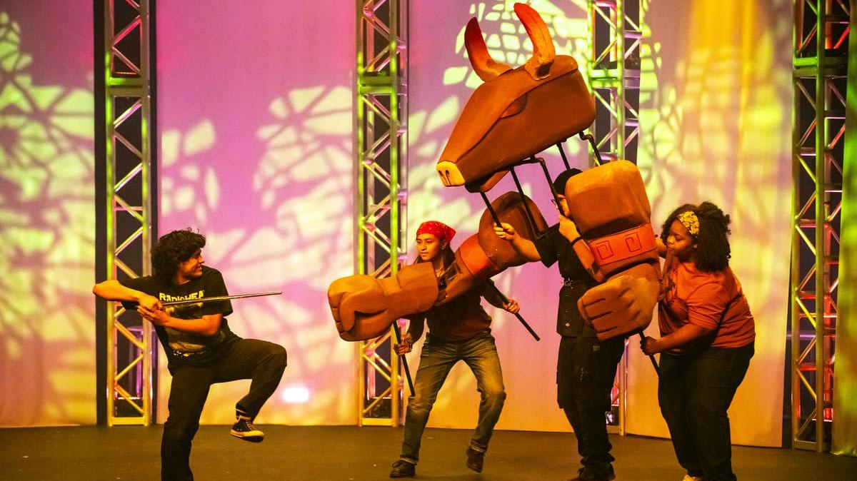 Performers on stage manipulate a large animal puppet during a theatrical scene.