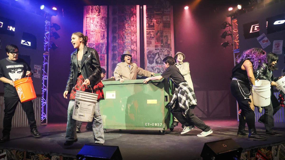 Stage performers push a large green dumpster while carrying buckets during a live theatrical performance.