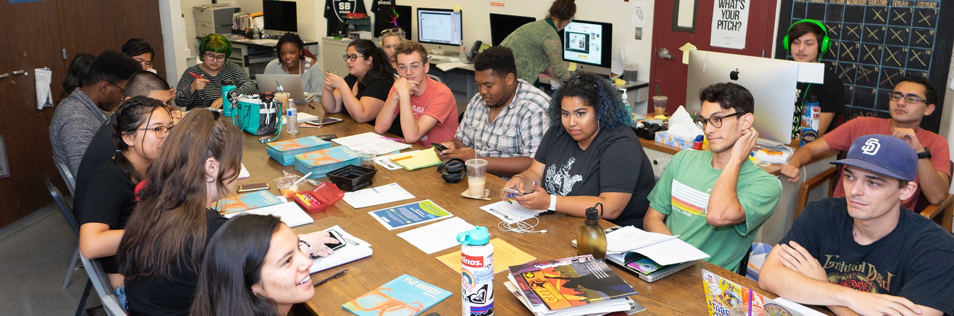 A group of diverse journalism students are gathered around a table.