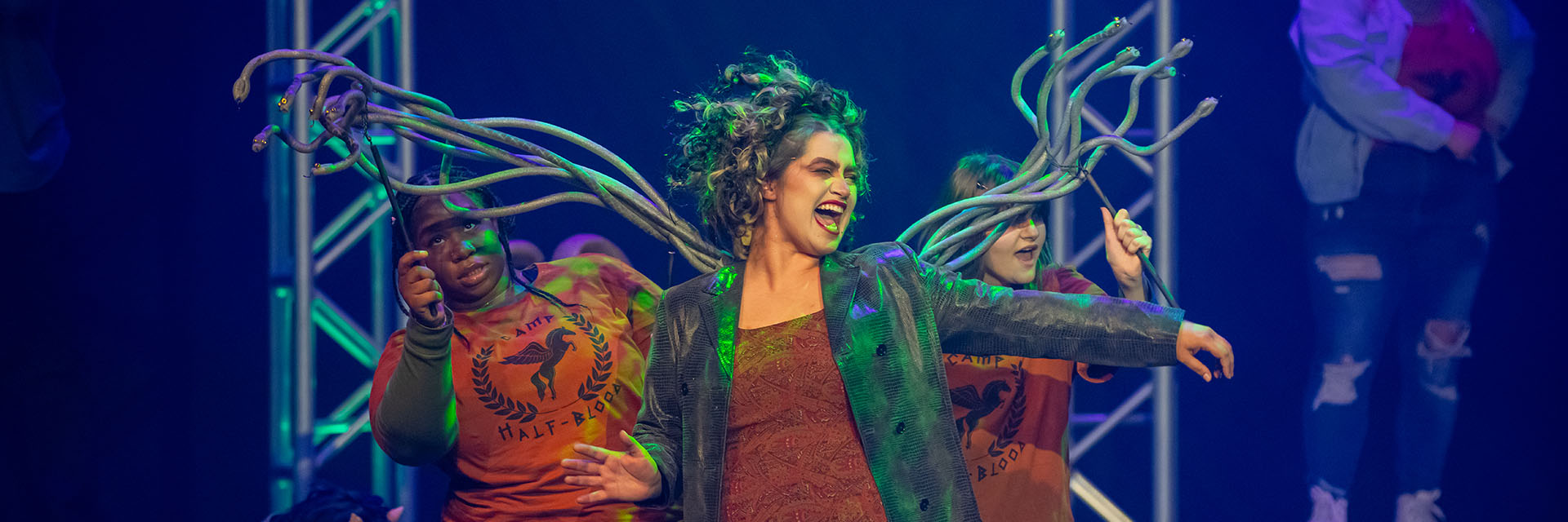 A lively stage performance features three actors. The central figure, embodying Medusa, laughs energetically alongside two actors in CAMP HALF-BLOOD shirts.