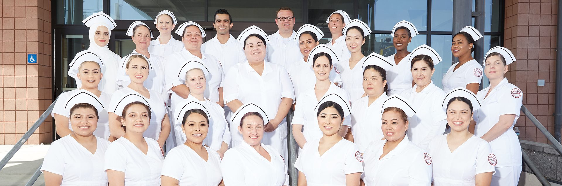 A diverse group of smiling nursing students in white uniforms and caps, posing outdoors.