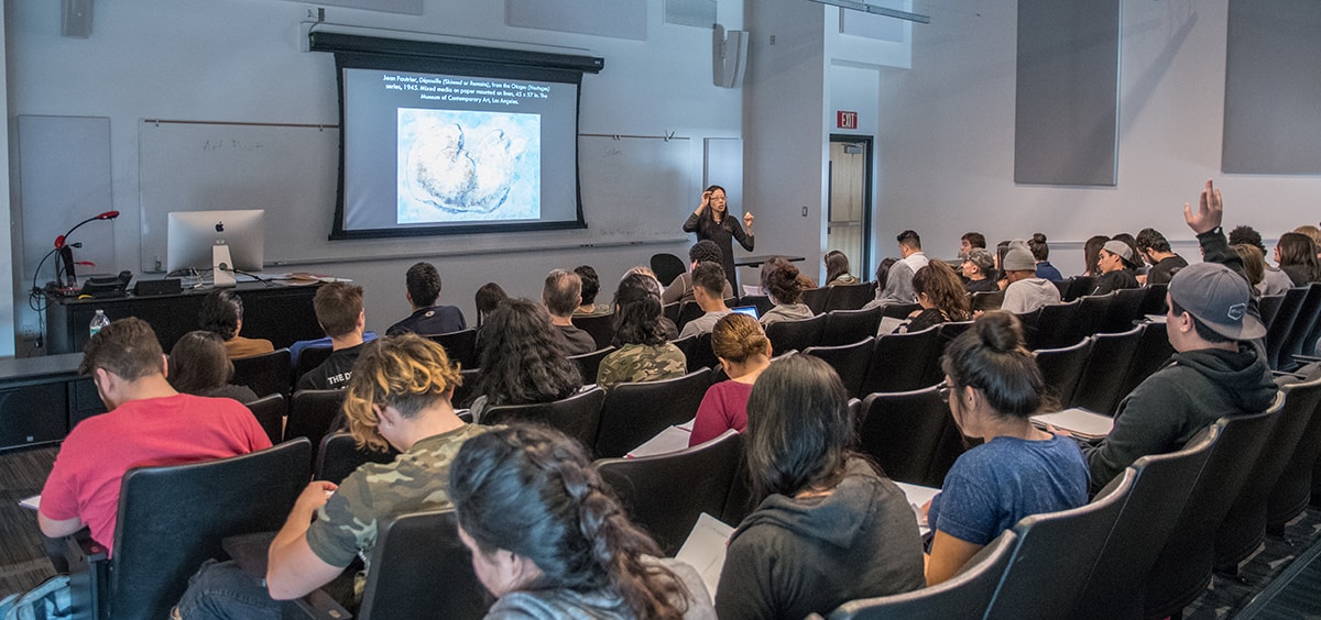 A professor lectures in a college classroom. Students sit in rows of seats facing a screen displaying artwork.