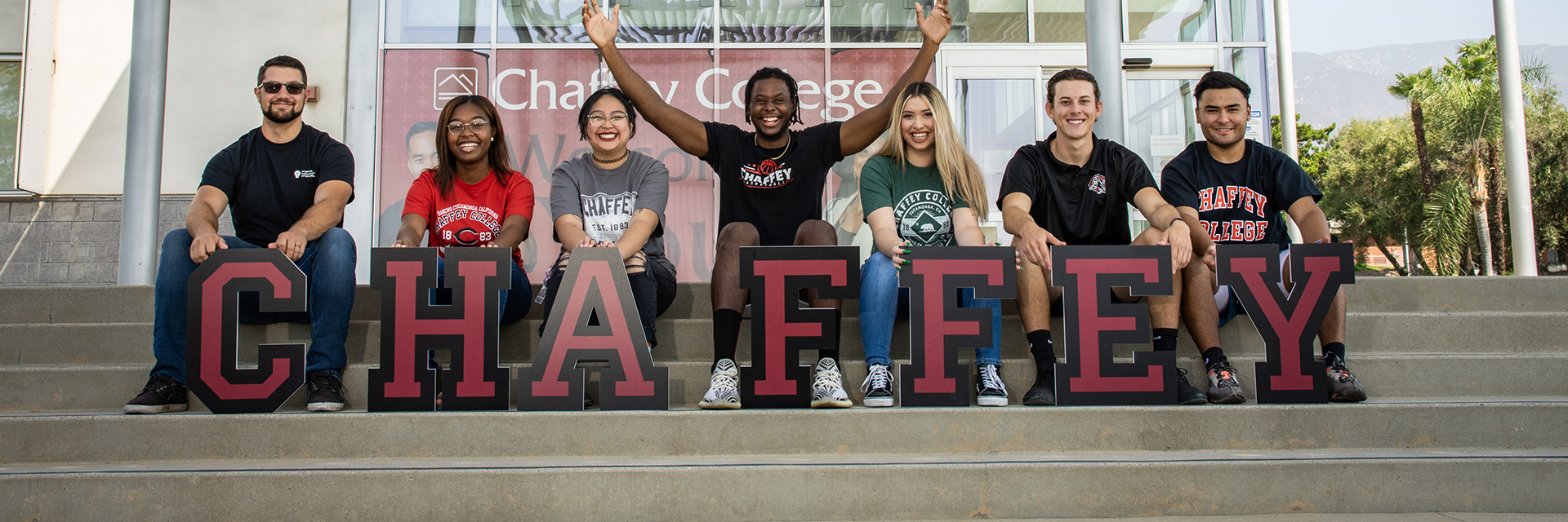 Seven smiling Chaffey College students sit on stairs holding up large maroon letters that spell CHAFFEY. The group is diverse, and mountains are visible in the background.