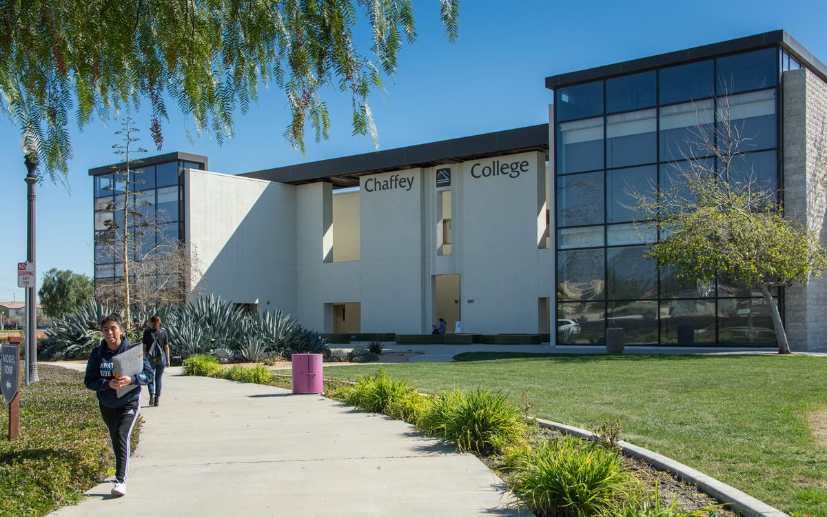 Students walk along a sunny pathway in front of the modern Chaffey College building, featuring large glass windows and surrounded by lush greenery.