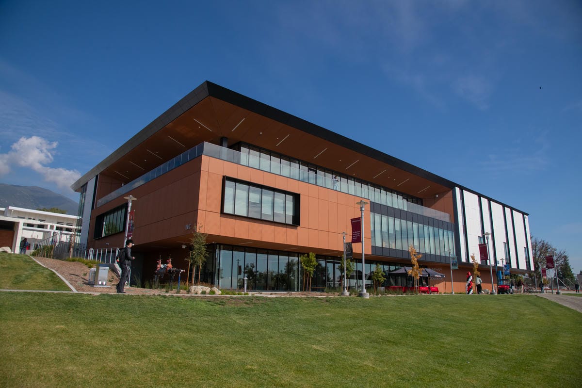 A modern, angular building with large glass windows and an orange facade stands against a clear blue sky. It is set on a grassy lawn with a few small trees and banners lining the walkway. A person walks along the path in front of the building.