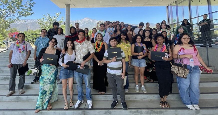 A large group of graduating students stand on stairs outdoors, some holding diplomas. The building is modern, with mountains visible in the background.