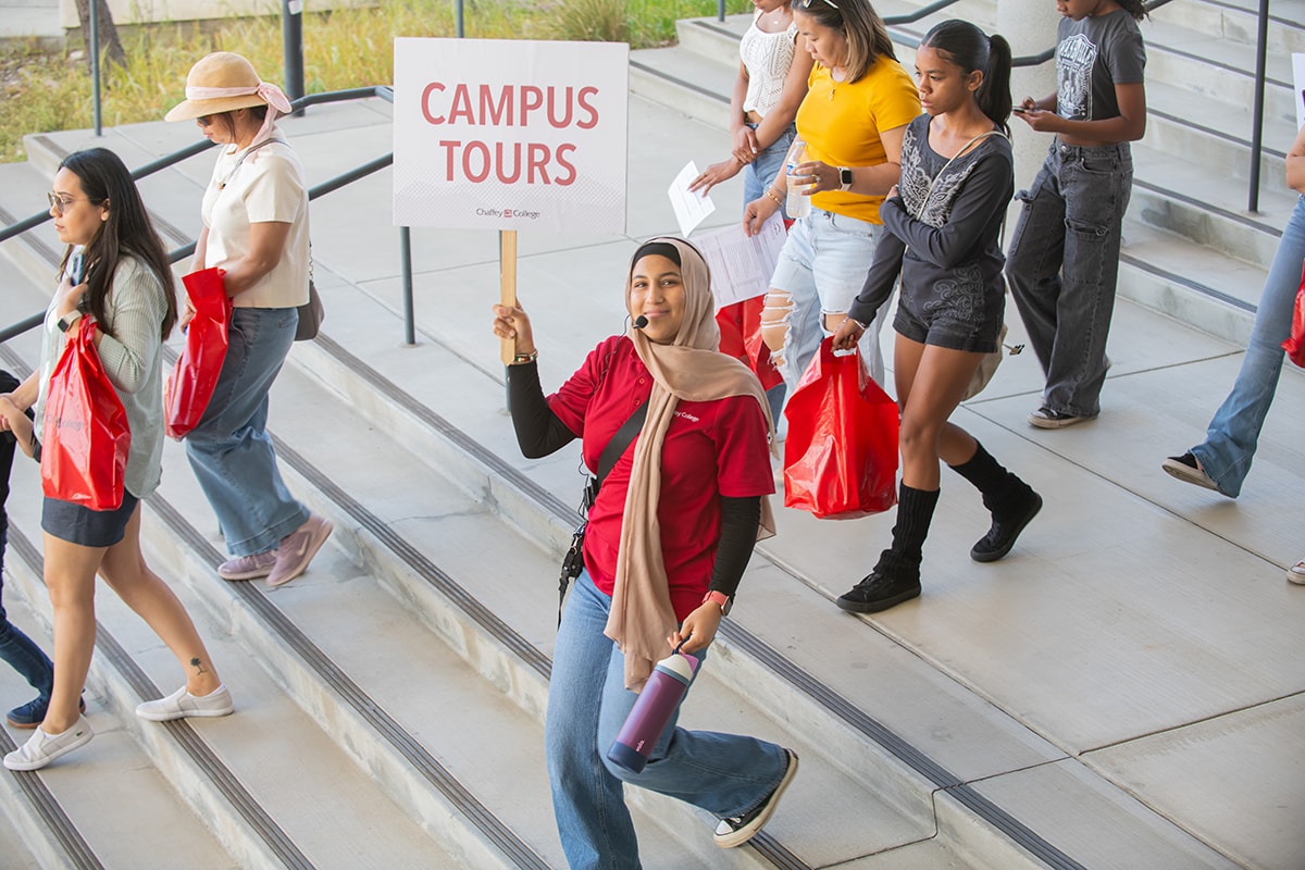 A student leads a campus tour with a sign that reads "CAMPUS TOURS", as prospective students follow. The tour guide is smiling and carrying a water bottle.