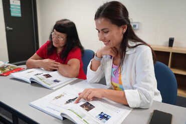 Two women are engaged in a shared learning activity with open books. The woman on the right points to an image in her book. Text reads New vocab?.