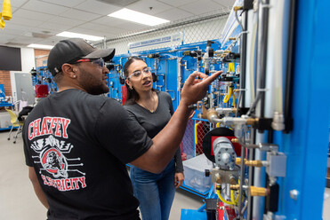 A man in a CHAFFEY ELECTRICITY shirt points to complex lab equipment as a woman observes. They are likely in a vocational training or classroom setting.