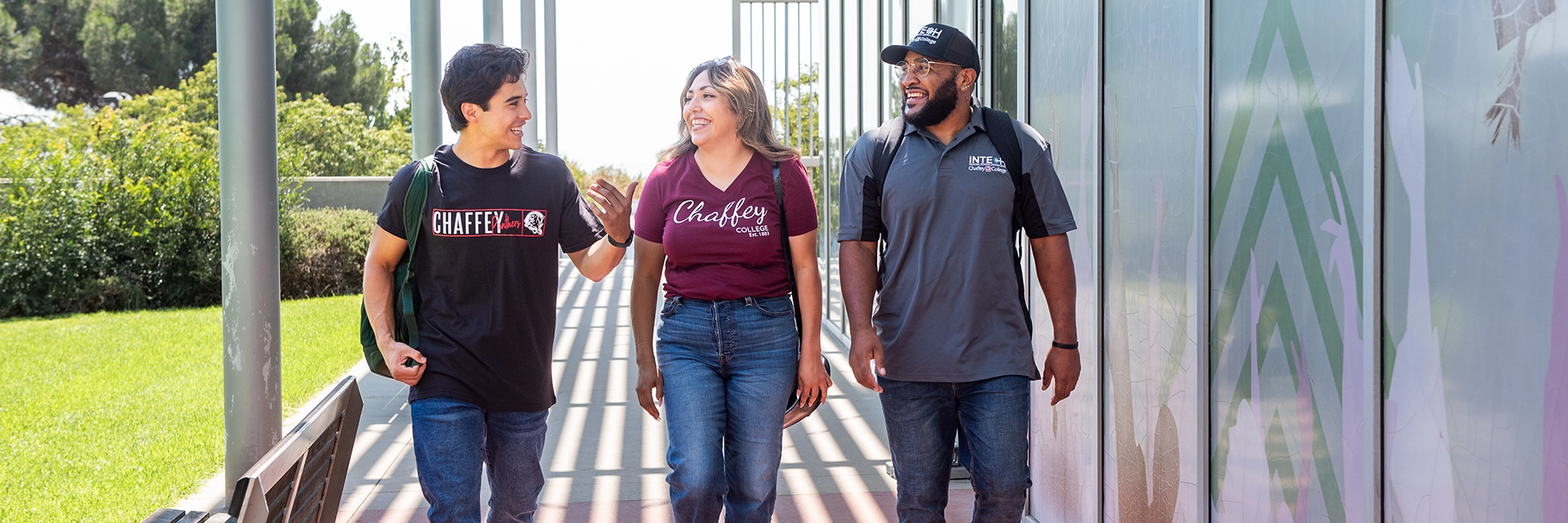 Three students walk together outside, smiling and chatting. They are wearing casual clothes with Chaffey College logos, and each carries a backpack. The setting is bright and sunny, with greenery in the background and patterned glass panels on one side.