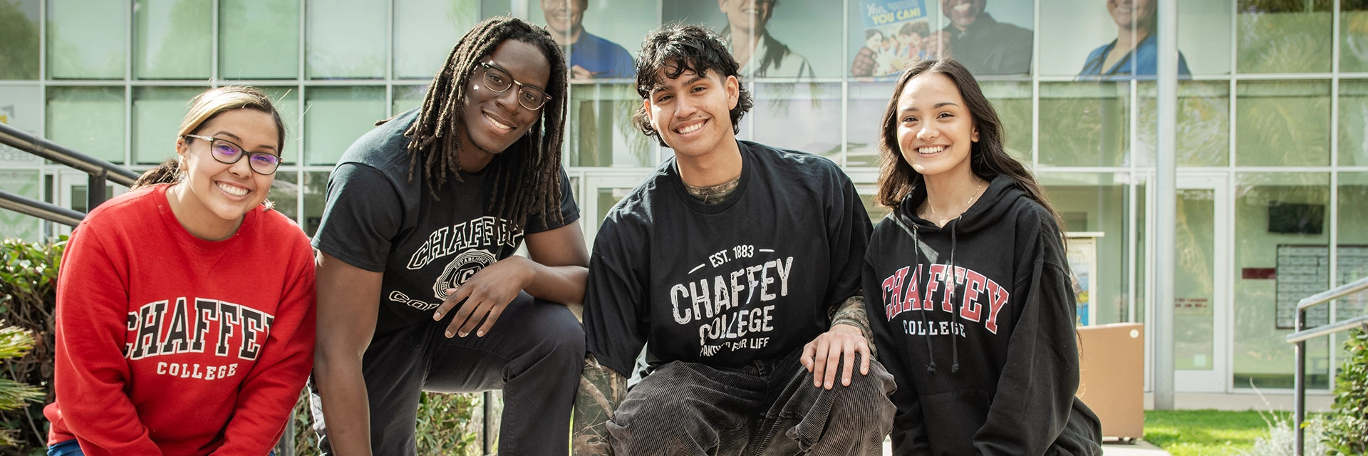 Four smiling students are wearing Chaffey College apparel. They are posing in front of a modern building with Chaffey College text on their clothing.