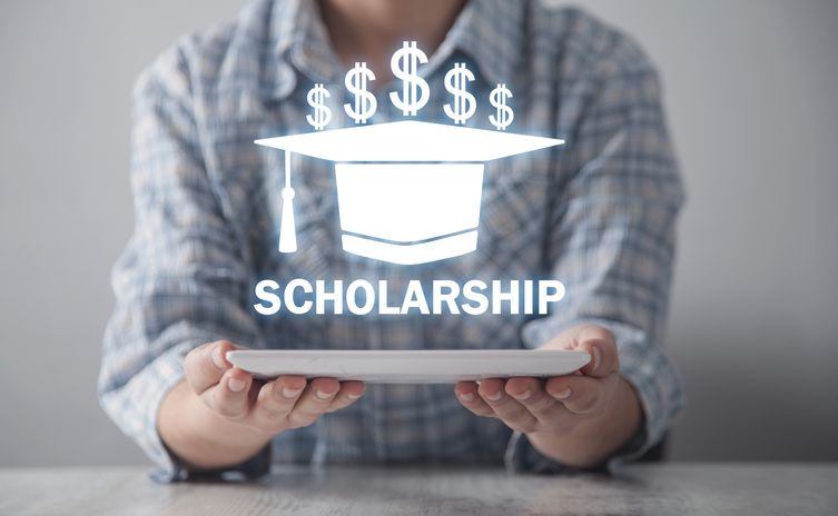 A person holds a tablet, with a glowing graphic above it showing a graduation cap, dollar signs, and the word SCHOLARSHIP.