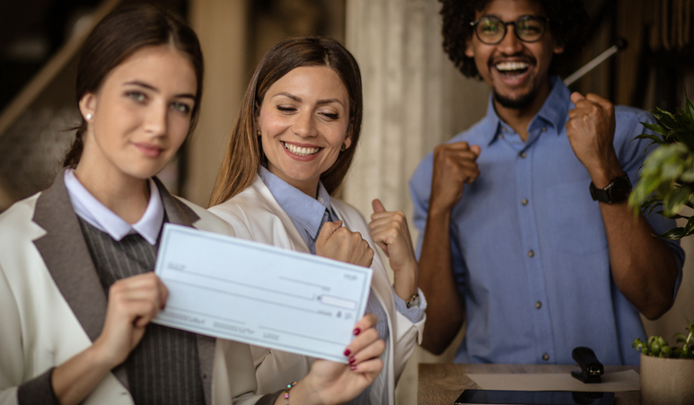 Three smiling colleagues celebrate, with one woman holding a blank check. Another woman and a man enthusiastically pump their fists in success.