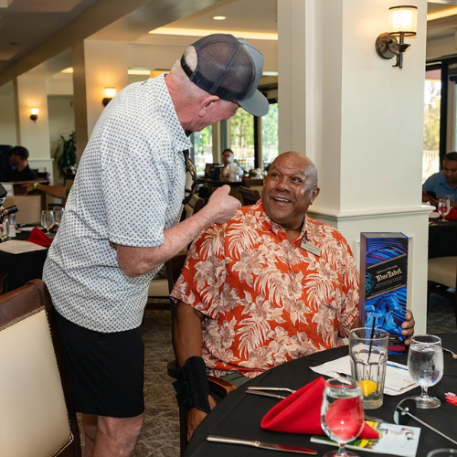 A man in a cap leans towards a smiling Chaffey College board member in a floral shirt at a table with a Blue Label bottle. They appear to be enjoying a social event.