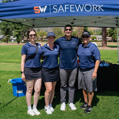 Four smiling people in matching navy shirts stand under a blue tent at an outdoor event. The tent displays SW SAFEWORK