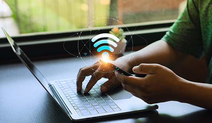 A person uses a laptop while also using a mobile phone. A Wi-Fi graphic overlay implies a strong internet connection.