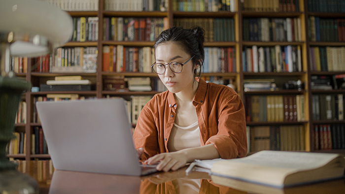 Focused young woman doing research in the library, typing an article on laptop