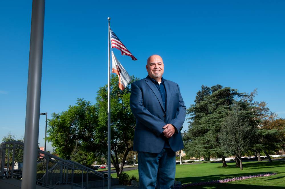 Alfredo Zendejas stands in front of the Chaffey admissions building. The American flag waves in the background.