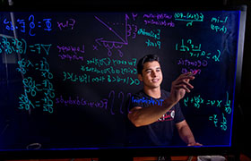 A student is writing complex mathematical equations on a transparent board, using colorful markers. The individual appears focused and is dressed casually, set against a dark background that emphasizes the vibrant writing.