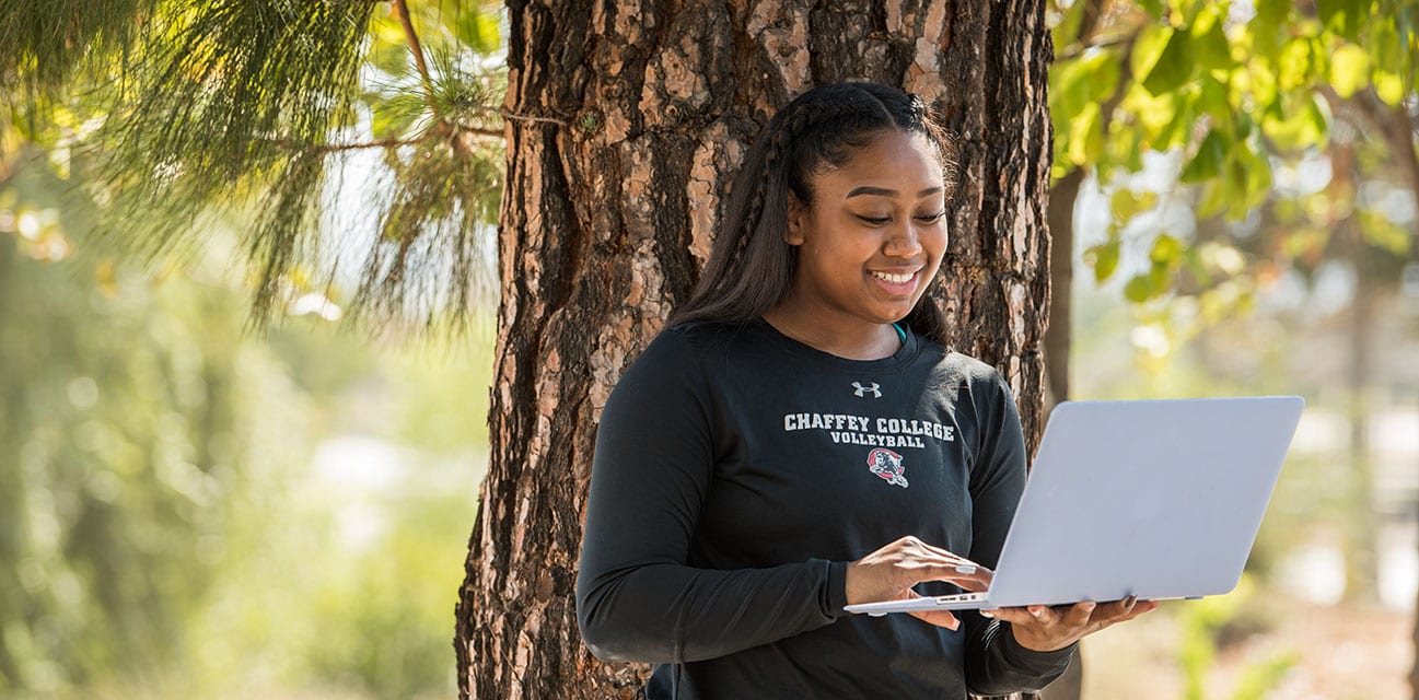 A student wearing a Chaffey College Volleyball shirt smiles while using a laptop, leaning against a tree in a sunlit outdoor setting.