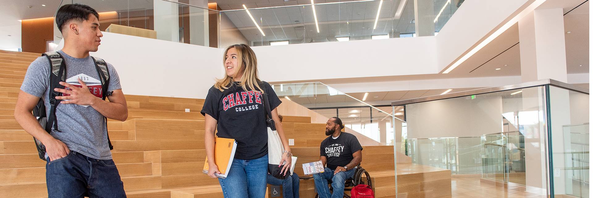 Students are in a modern college building with large wooden steps. A student in a CHAFFEY COLLEGE shirt holds books, while another sits in a wheelchair nearby, also wearing CHAFFEY COLLEGE.