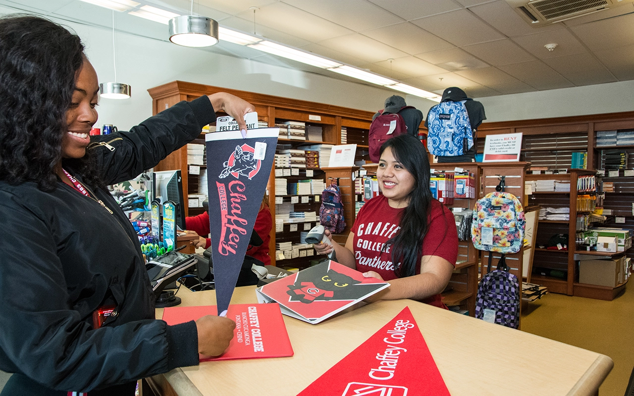Two smiling employees stand at a Chaffey College store counter. Merchandise with the college's logo is displayed; one pennant reads Chaffey College Panthers.