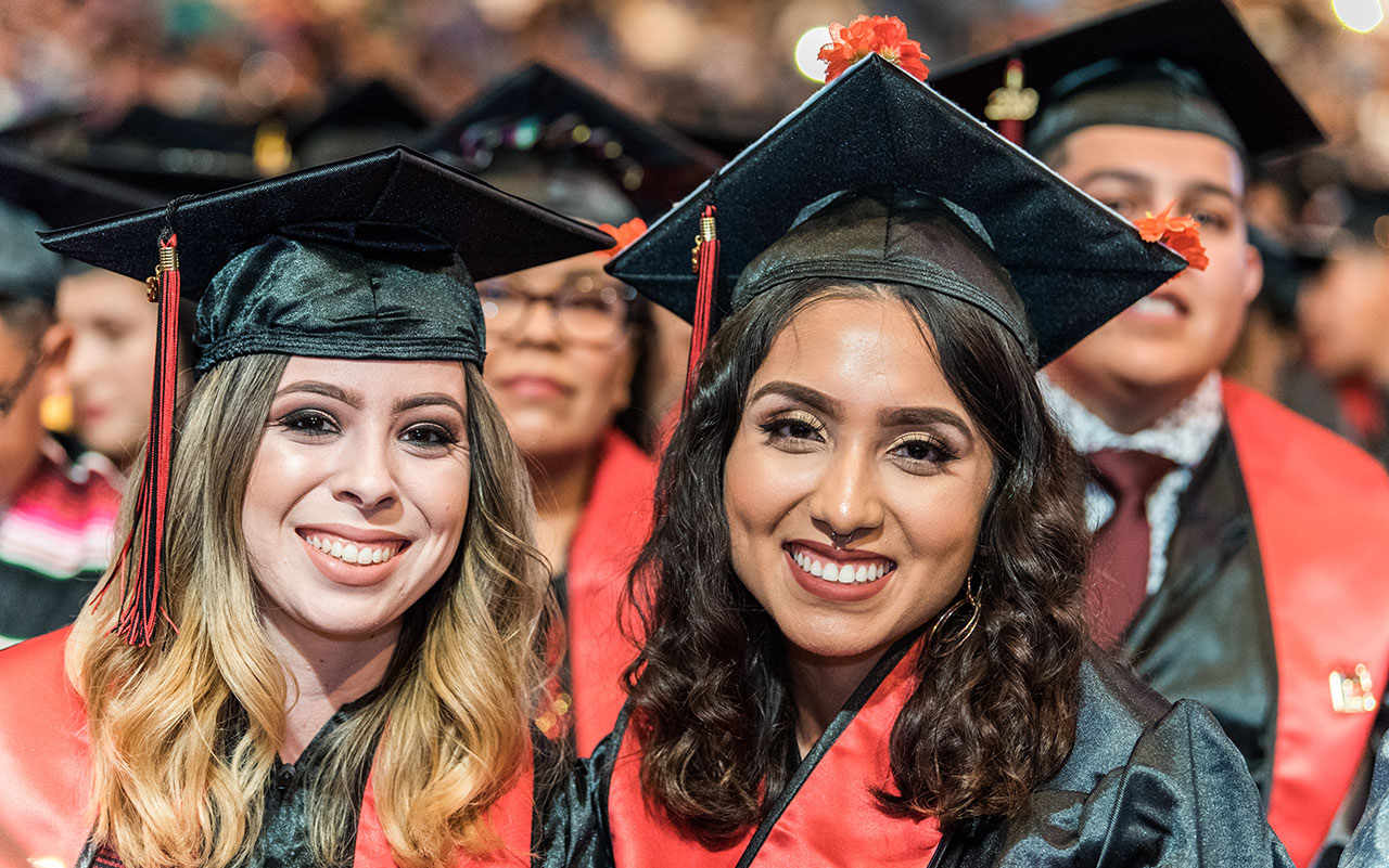 Two smiling graduates wearing black caps with red tassels and red stoles pose together at a ceremony, surrounded by other graduates in the background.
