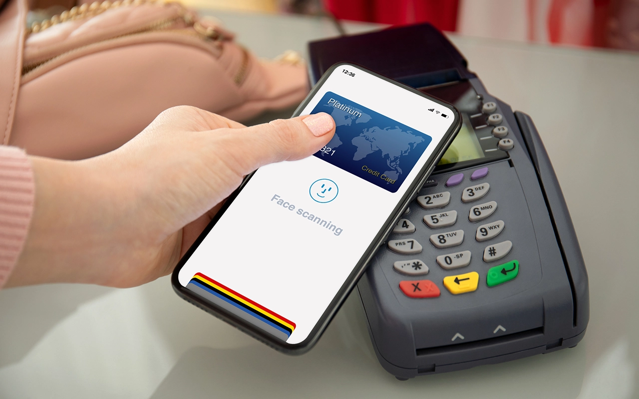 A person uses their smartphone to pay at a terminal. The phone displays a platinum credit card and the words Face scanning.