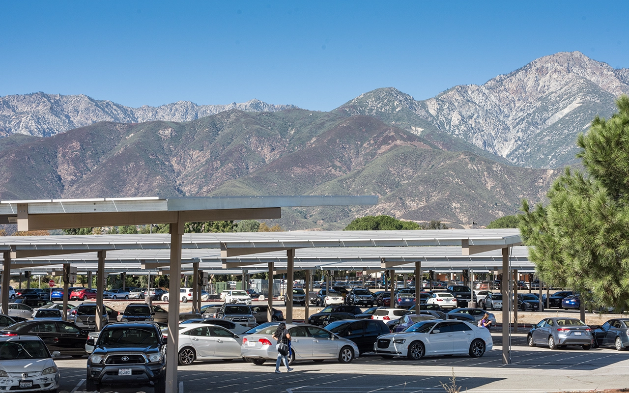 A parking lot with solar panel shade structures is shown with a backdrop of mountains. Many cars of varied color and model fill the lot on a clear sunny day.